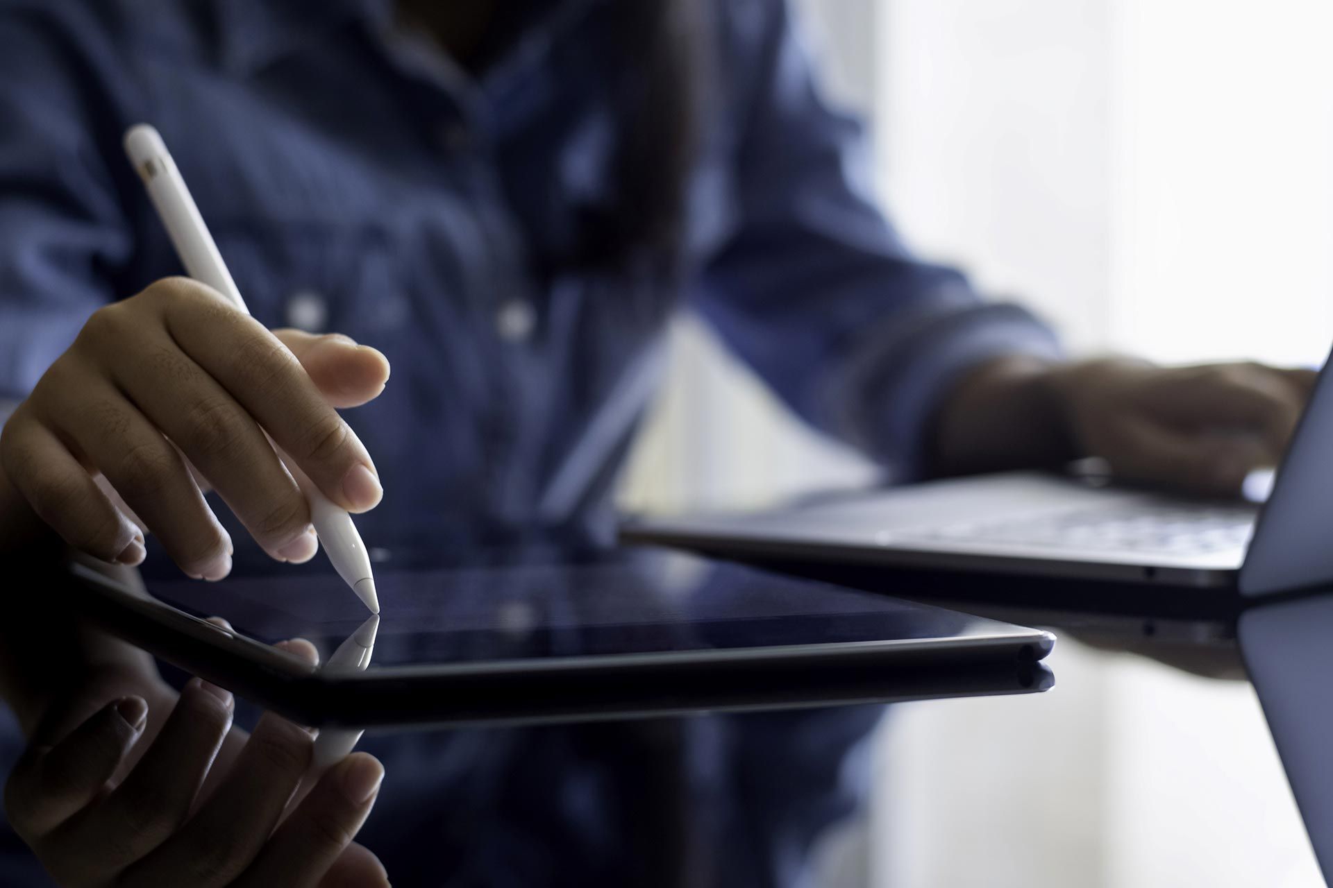 Eine Person arbeitet parallel mit einem Tablet-Computer und einem Notebook. Das Bild ist in Blautönen gehalten und der gezeigte Mensch trägt ein dunkelblaues Hemd. Der Fokus des Fotos liegt auf den Händen der Person.